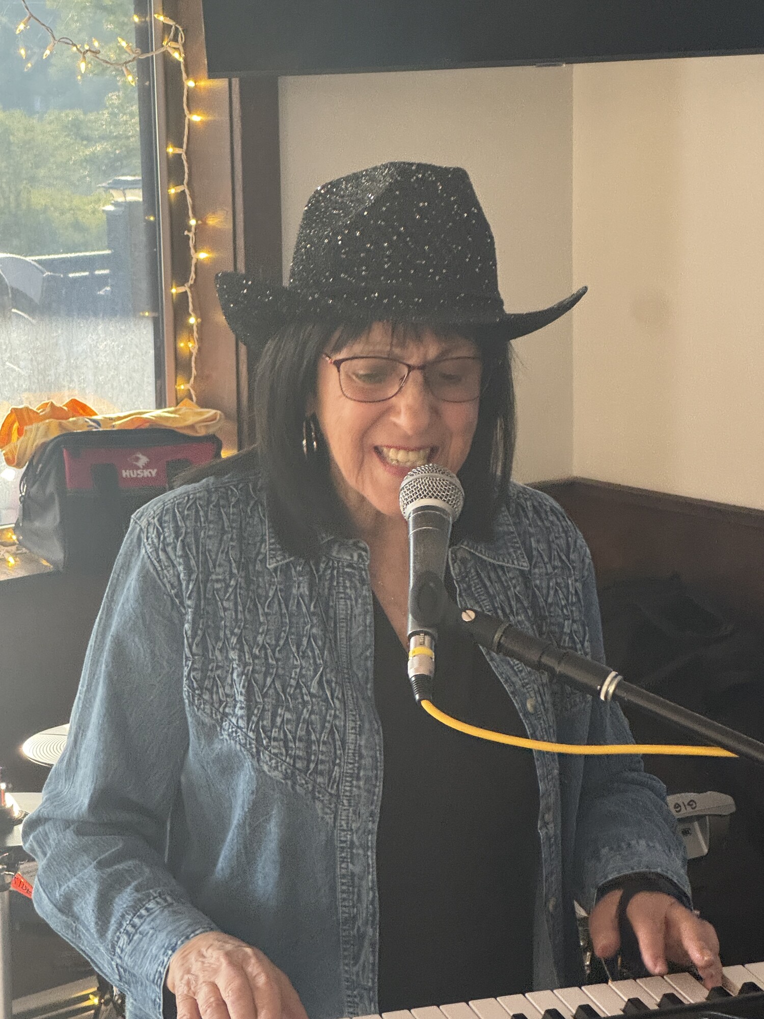 Beverly singing at the keyboard in a black sparkly hat and denim jacket
