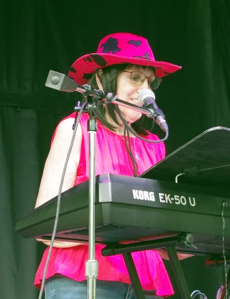 Beverly singing at the keyboard in a pink hat on an outdoor stage